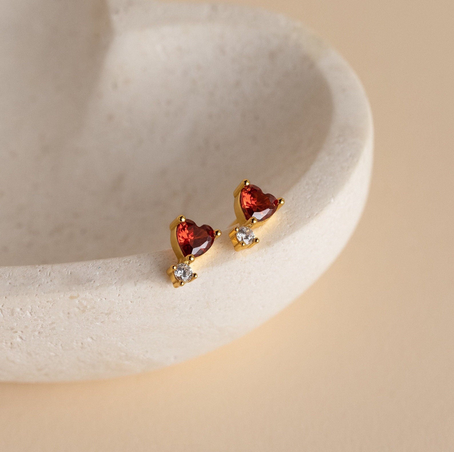 Garnet Heart Studs: gold stud earrings with heart-shaped garnet stones and sparkling clear gems, displayed on a textured cream dish.