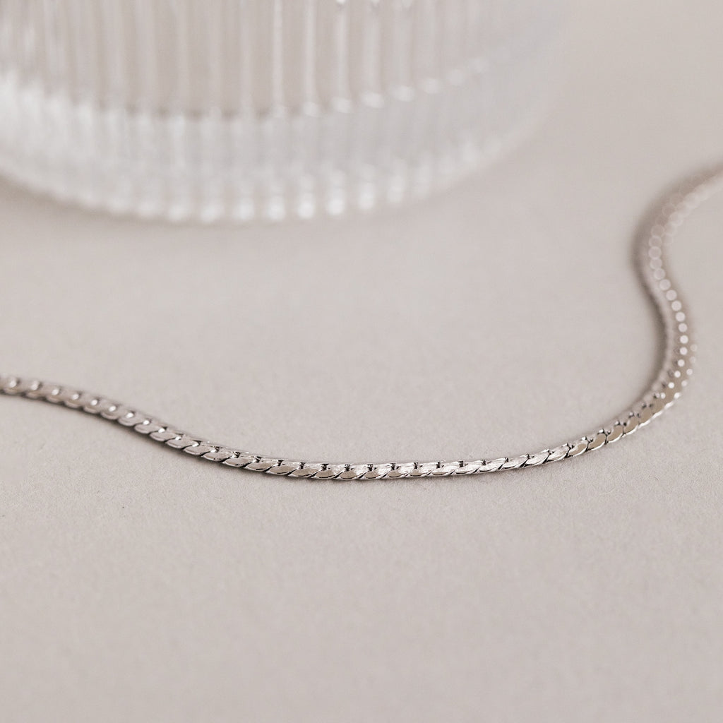 A minimalist necklace, this Men's Snake Chain Necklace is displayed on a light surface beside a textured glass object.