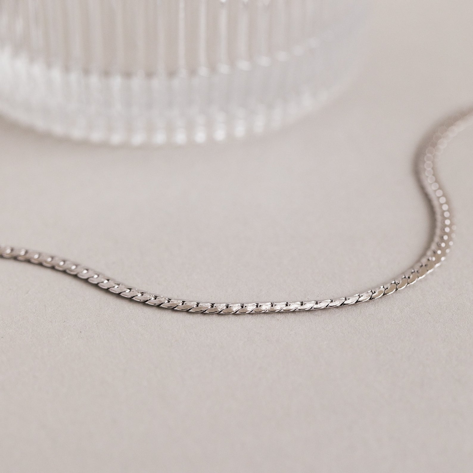 A minimalist necklace, this Men's Snake Chain Necklace is displayed on a light surface beside a textured glass object.
