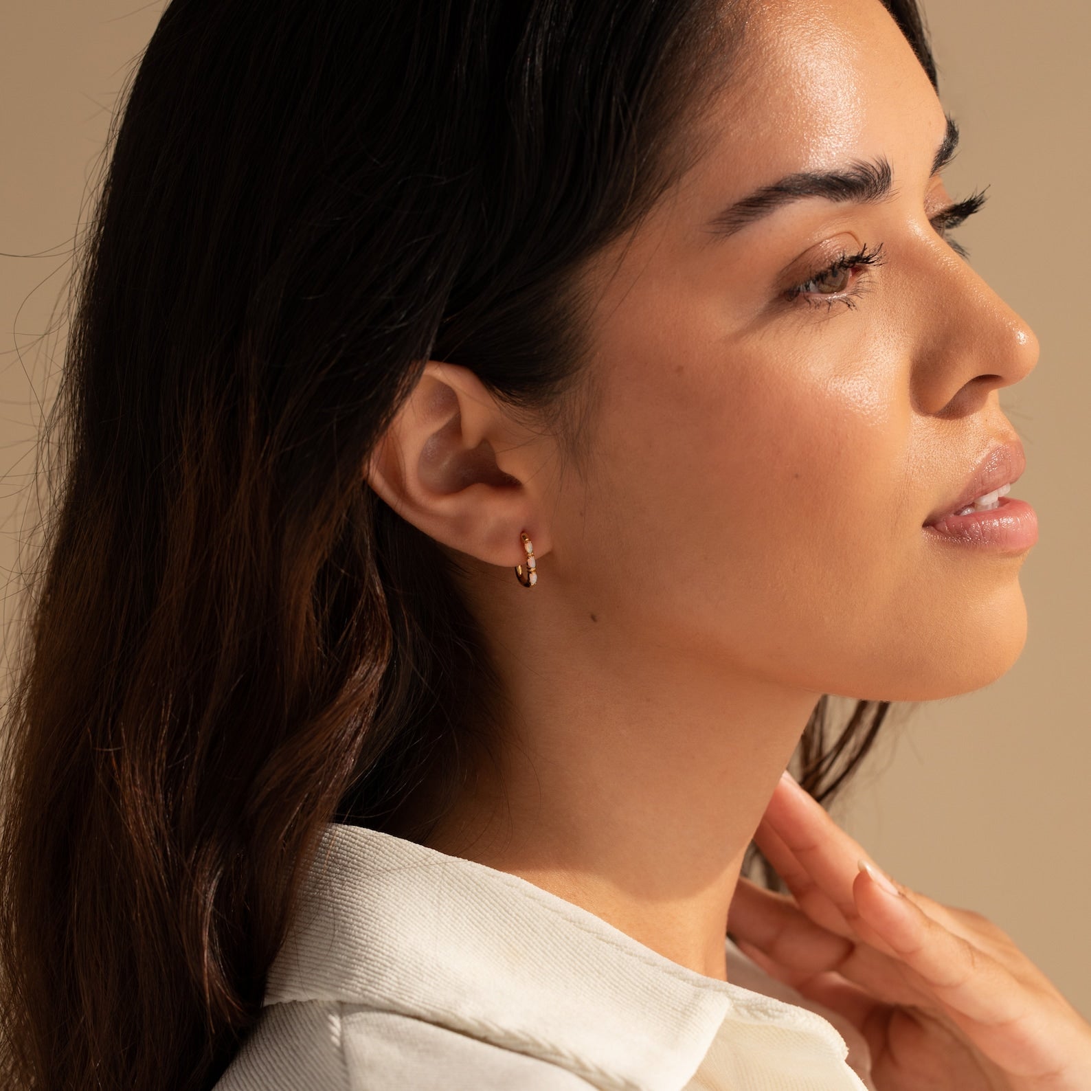 Woman with long dark hair looks to the side in soft, warm lighting, wearing Halo Opal Hoops—elegant gold huggie earrings.