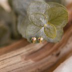 Mushroom Studs: Gold and purple earrings adorned with CZ diamonds, displayed on green leaves over wood—a charming, nature-inspired jewelry piece.