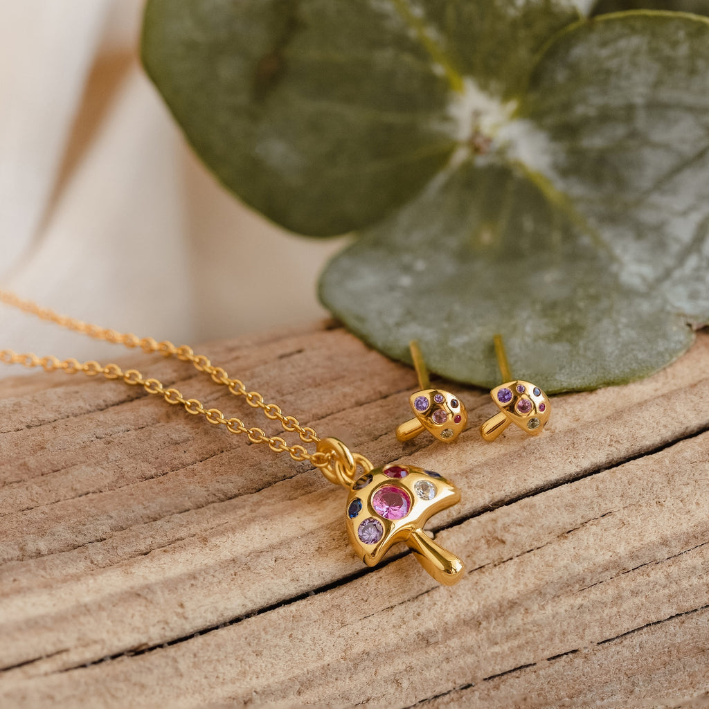 Mushroom Studs, gold mushroom-shaped earrings adorned with colorful gems and CZ diamonds, rest on wood with green leaves in the background—a perfect piece of nature-inspired jewelry.