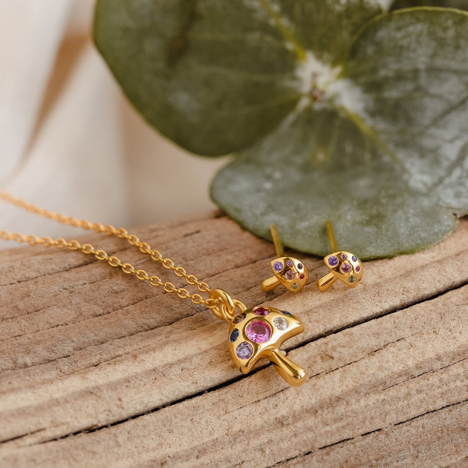 Mushroom Studs, gold mushroom-shaped earrings adorned with colorful gems and CZ diamonds, rest on wood with green leaves in the background—a perfect piece of nature-inspired jewelry.