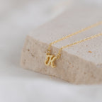 The Mellow Dainty Initial Necklace features a delicate K pendant, perfect as a custom accessory, shown elegantly resting on a light stone surface.