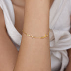 The Tiny Multiple Name Bracelet features customizable cursive charms, shown on a wrist with minimal jewelry and a white sleeveless top.