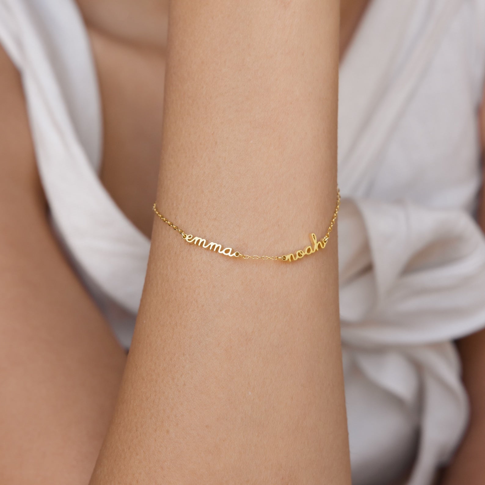The Tiny Multiple Name Bracelet features customizable cursive charms, shown on a wrist with minimal jewelry and a white sleeveless top.