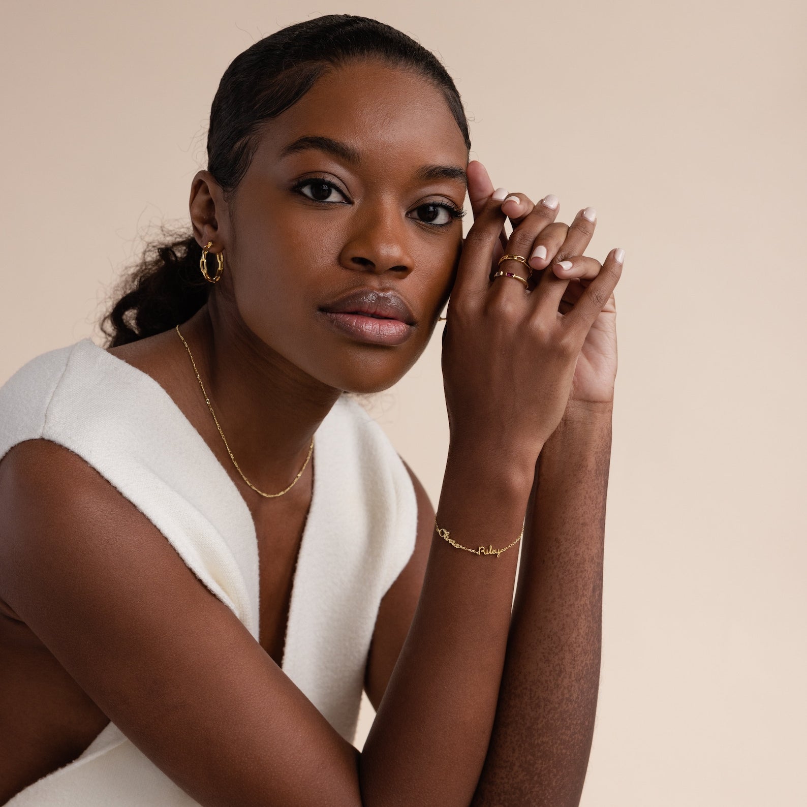 A woman in a white sleeveless top wears minimal jewelry—a gold Tiny Multiple Name Bracelet—as she rests her hands near her face against a neutral background.