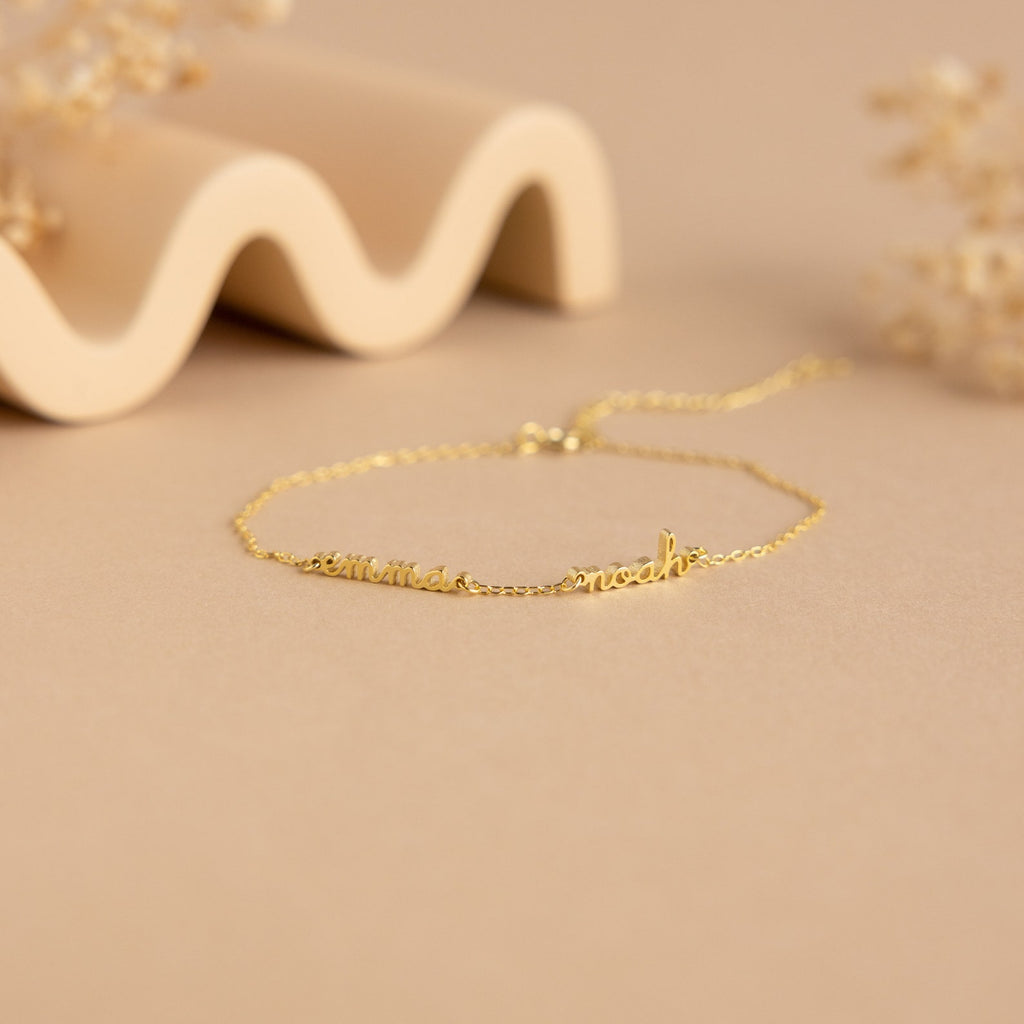 The Tiny Multiple Name Bracelet features a delicate gold chain with customizable names in cursive, displayed on a beige surface—ideal for fans of minimal, personalized jewelry and unique name bracelet styles.