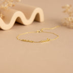 The Tiny Multiple Name Bracelet features a delicate gold chain with customizable names in cursive, displayed on a beige surface—ideal for fans of minimal, personalized jewelry and unique name bracelet styles.