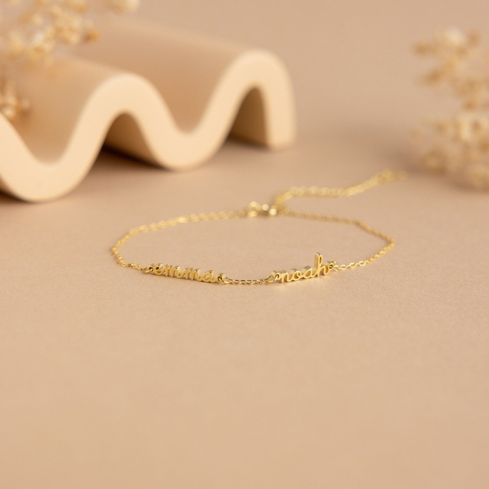 The Tiny Multiple Name Bracelet features a delicate gold chain with customizable names in cursive, displayed on a beige surface—ideal for fans of minimal, personalized jewelry and unique name bracelet styles.