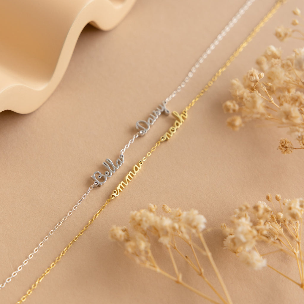 Gold and silver nameplate necklaces rest on a beige surface with dried flowers and a wavy accent, ideal for minimalists or anyone looking to pair them with the customizable Tiny Multiple Name Bracelet for a unique, personalized style.