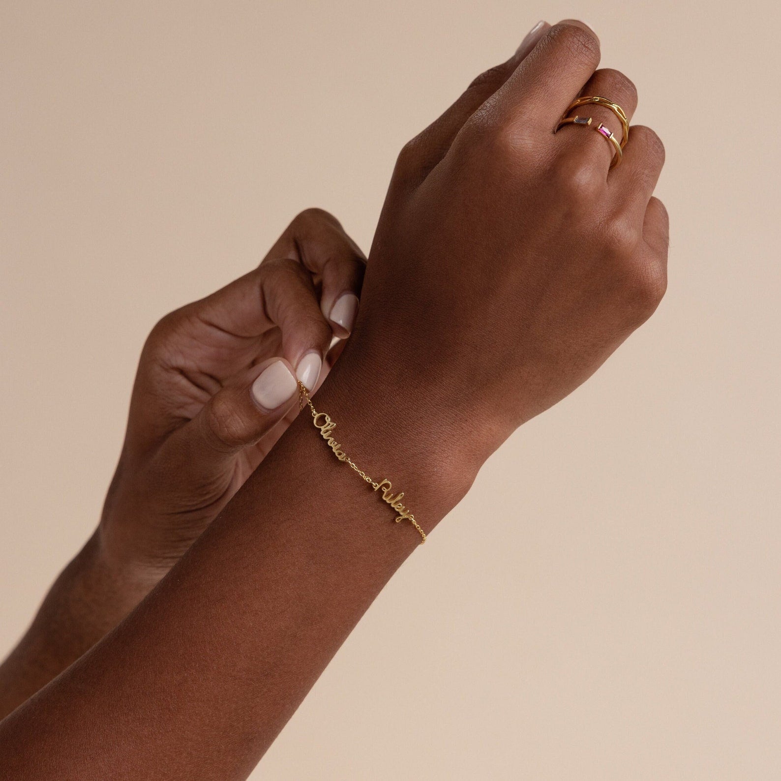 A person wears a Tiny Multiple Name Bracelet and gold rings, showcasing minimal jewelry against a beige background.