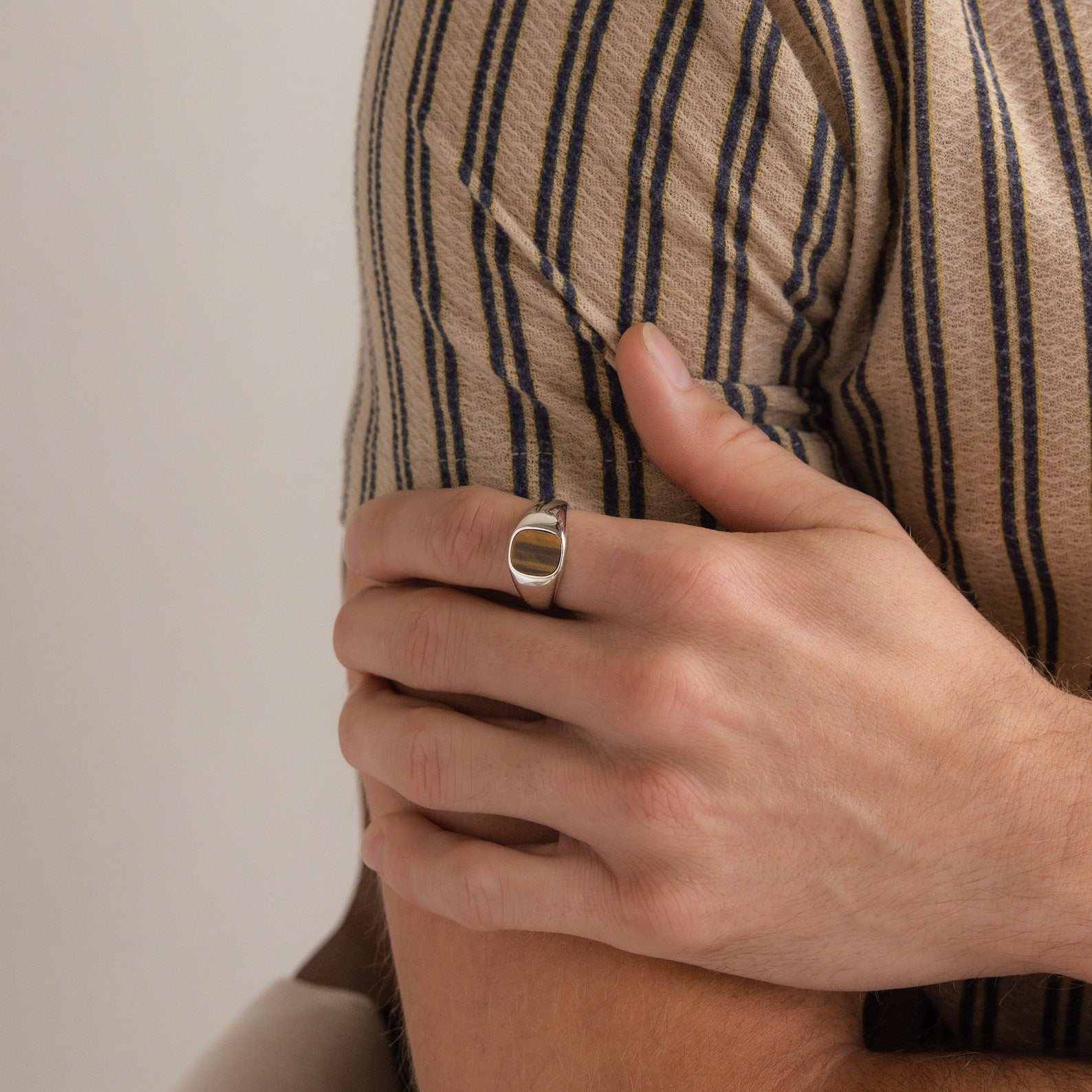 A person wearing a striped shirt gently rests their hand on their upper arm, showcasing the Tigers Eye Signet Ring in Sterling Silver.