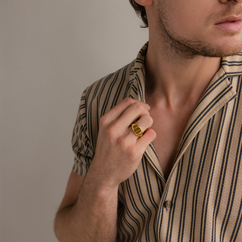 A man in a beige striped shirt holds his collar, displaying a bold Tigers Eye Signet Ring on his hand.