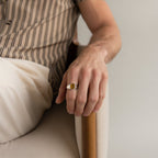 A person wearing a Tigers Eye Signet Ring sits on a beige chair with their hand resting on the armrest.