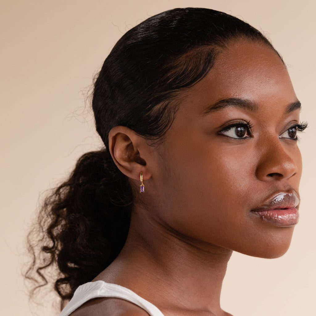 Model wearing a gold huggie earring with a baguette-cut amethyst charm, styled with a sleek ponytail and white tank top against a soft beige background.