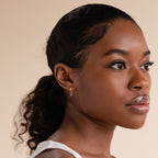 Model wearing a gold huggie earring with a baguette-cut amethyst charm, styled with a sleek ponytail and white tank top against a soft beige background.