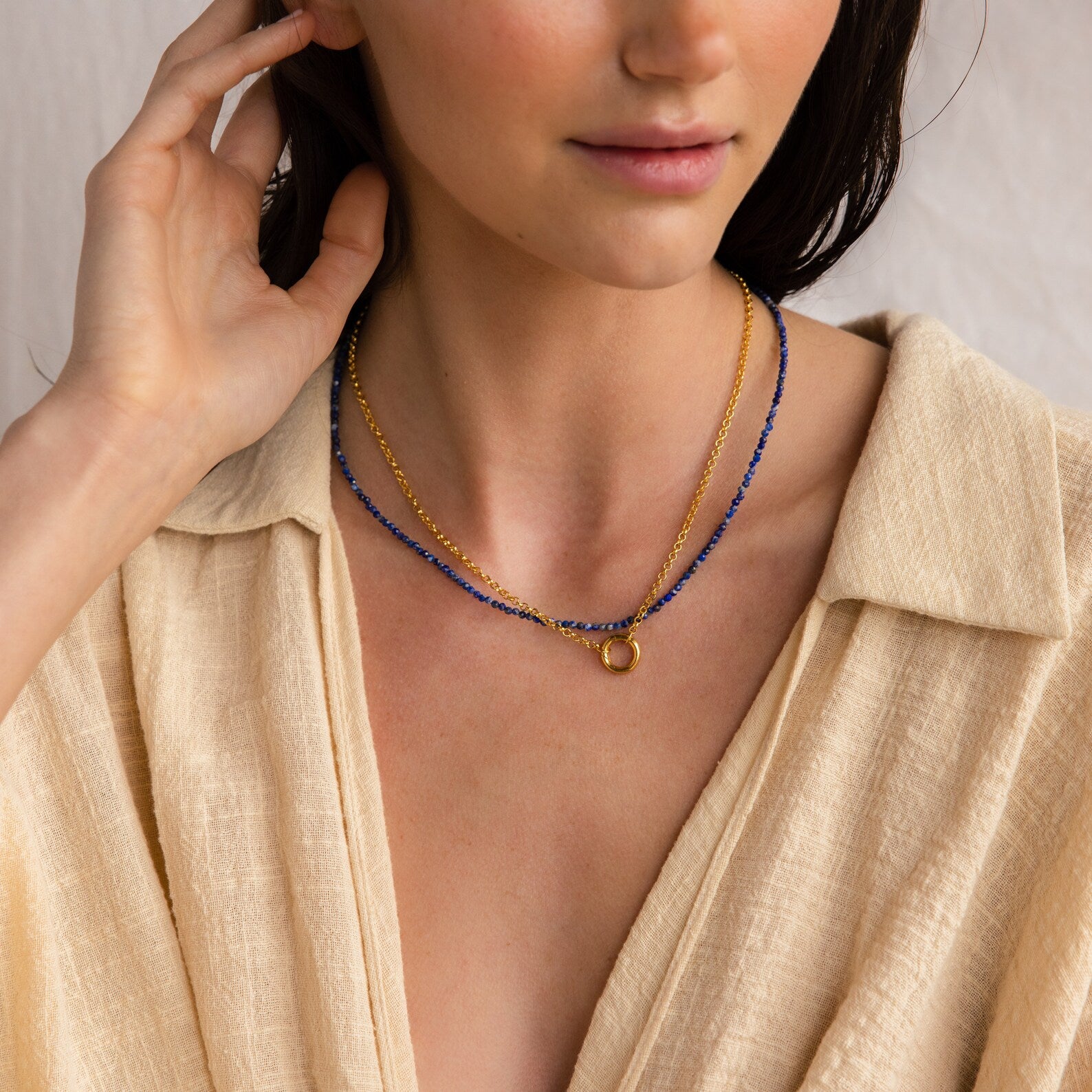 Woman in a beige shirt, shown from shoulders up, wears the Rolo Circle Charm Necklace in 18K Gold and layered gold and blue necklaces, touching her hair.