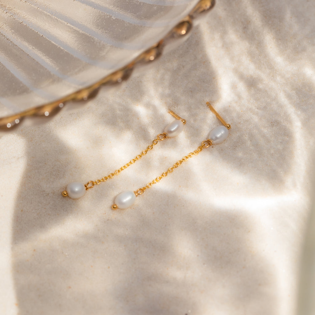 Pearl Drop Chain Earrings featuring white pearls are displayed on sunlit sand next to a glass dish, emphasizing the delicate elegance of this accessory.