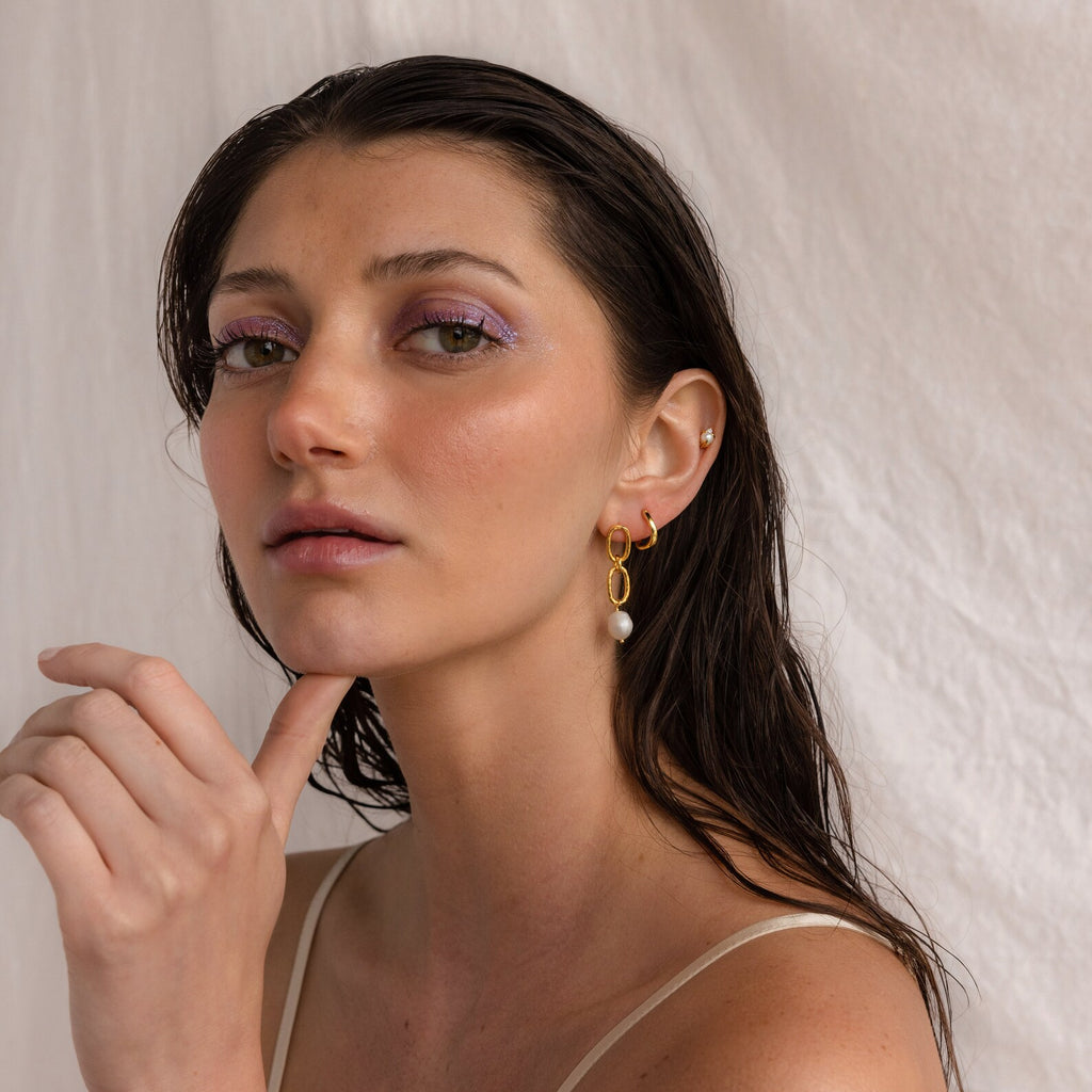 Woman with wet hair, wearing Pearl Drop Link Studs and lavender eyeshadow, poses against a light fabric background.