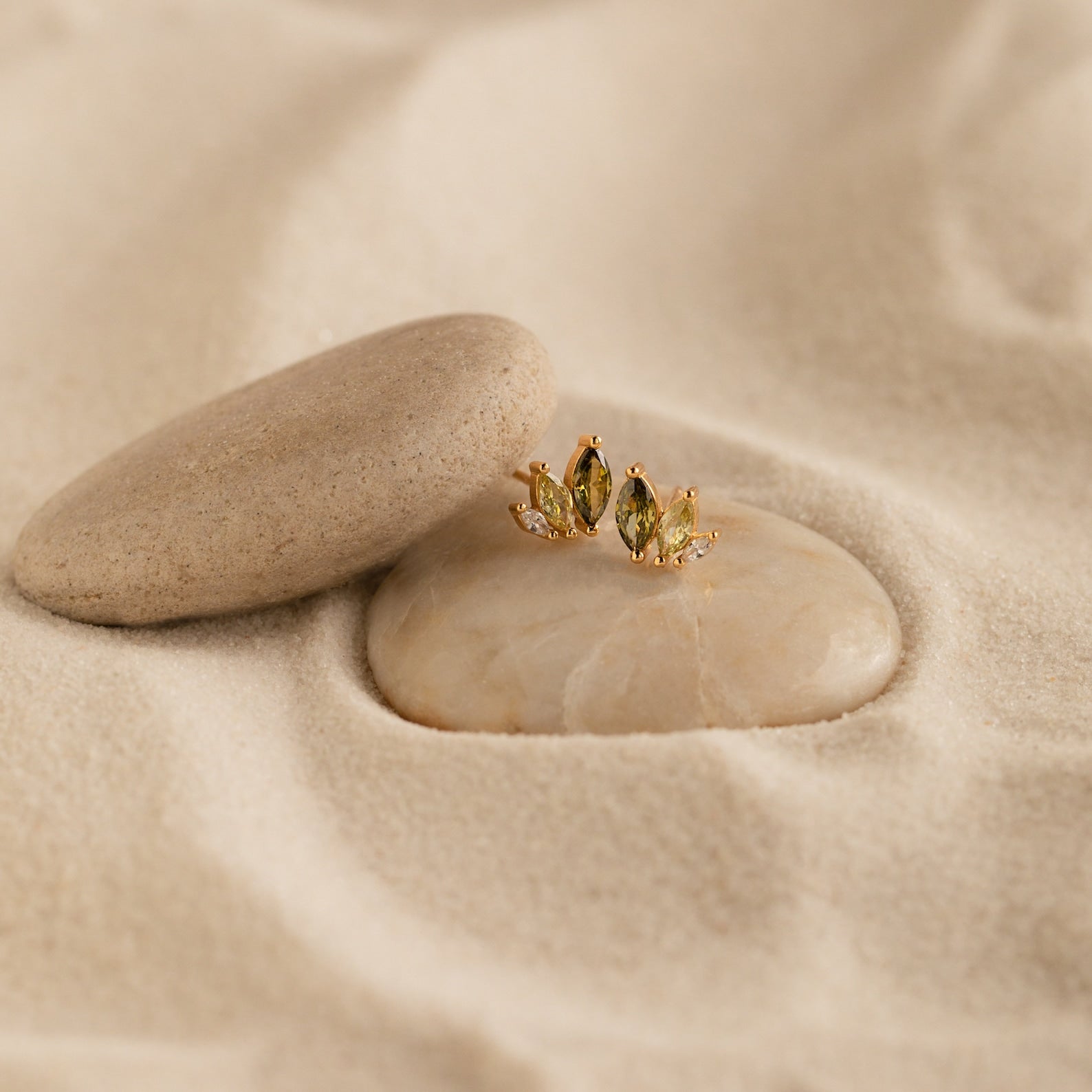 Seagrass Peridot Marquise Studs showcased on smooth stones nestled in soft sand.