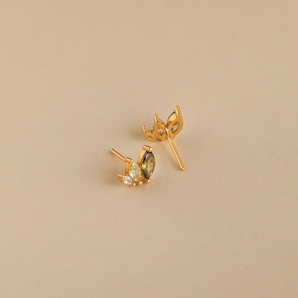 Seagrass Peridot Marquise Studs: Gold stud earrings with marquise and round-cut peridot stones add elegant sparkle against a beige backdrop.