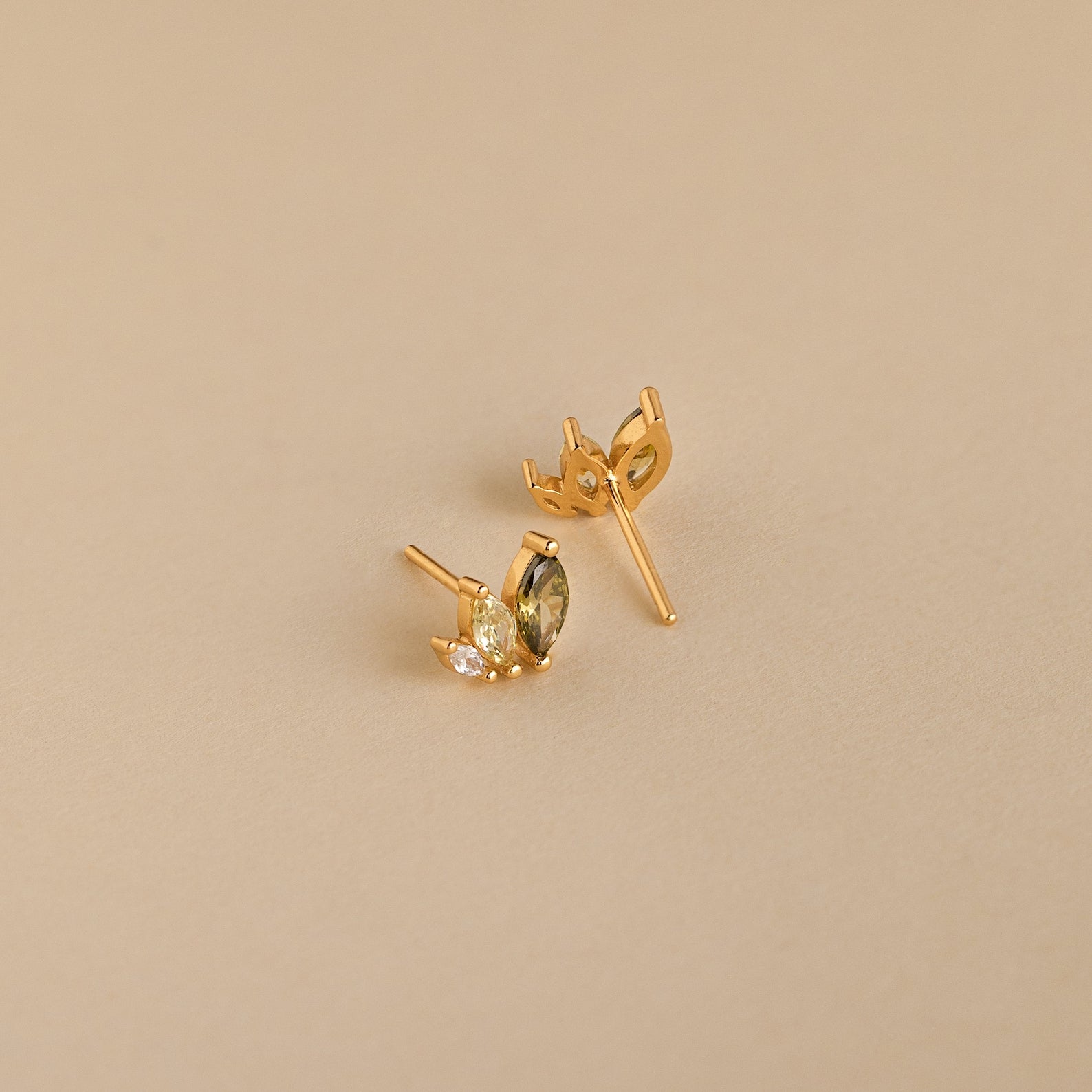 Seagrass Peridot Marquise Studs: Gold stud earrings with marquise and round-cut peridot stones add elegant sparkle against a beige backdrop.