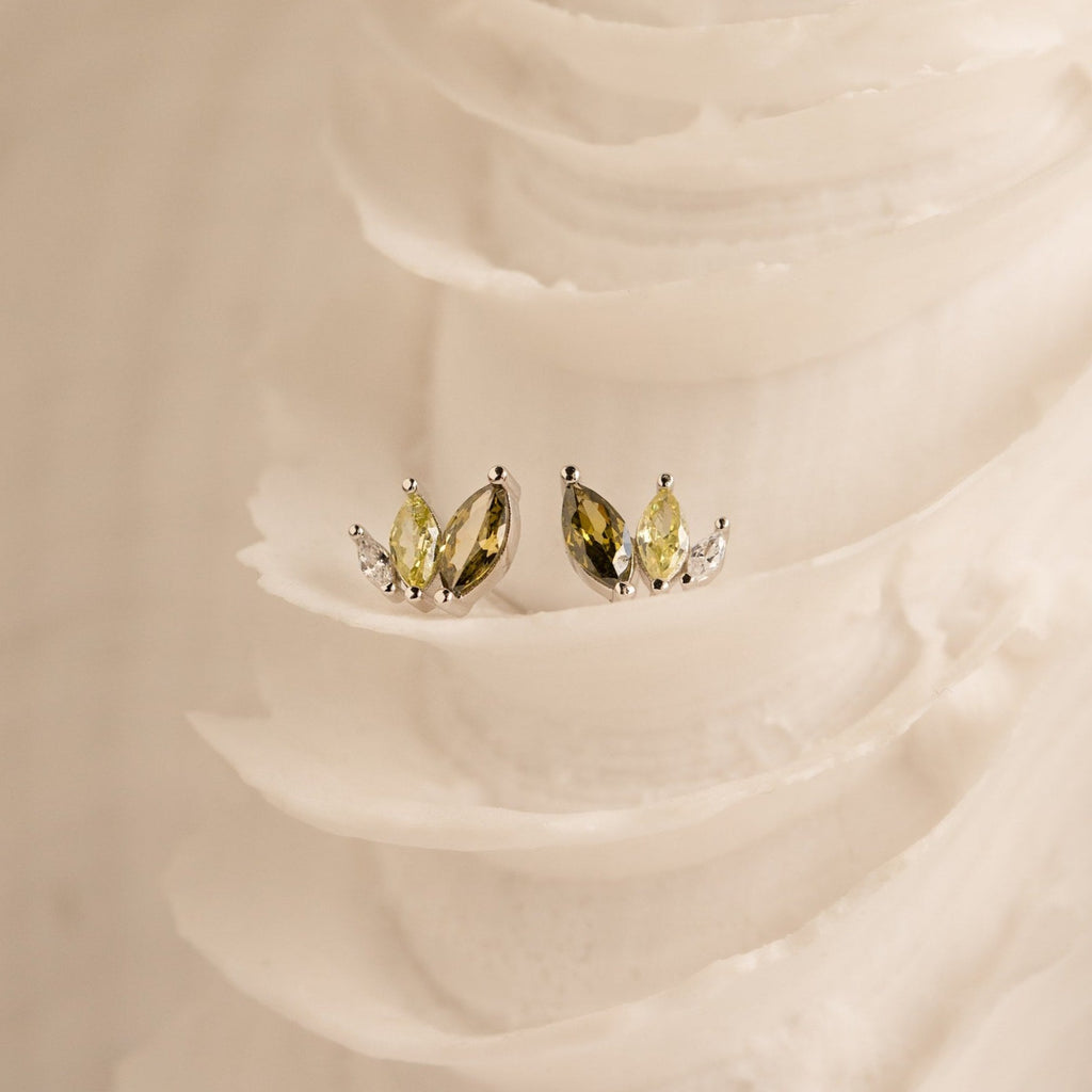 Gold and silver gemstone studs—including ombre marquise earrings and Seagrass Peridot Marquise Studs—are displayed on a textured, ivory-colored surface.