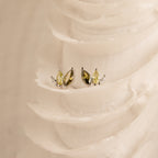 Gold and silver gemstone studs—including ombre marquise earrings and Seagrass Peridot Marquise Studs—are displayed on a textured, ivory-colored surface.