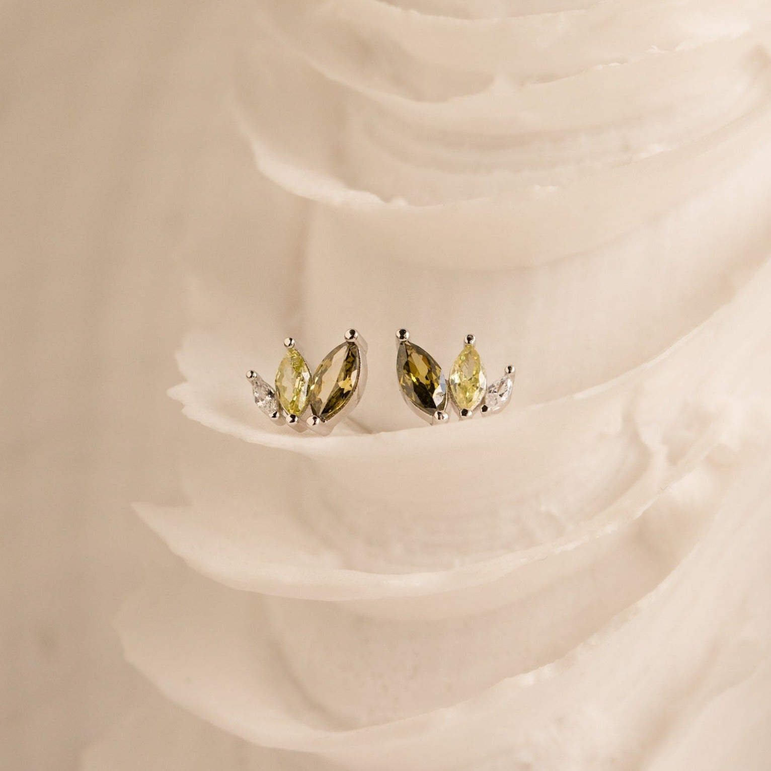 Gold and silver gemstone studs—including ombre marquise earrings and Seagrass Peridot Marquise Studs—are displayed on a textured, ivory-colored surface.