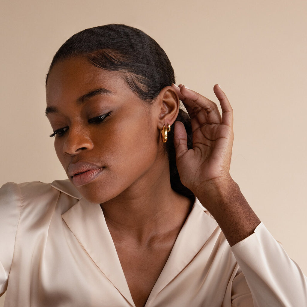 Woman in a cream blouse wears the Katrina Ribbed Hoops, oval earrings with a chunky ribbed texture, touching her ear against a beige background and embodying modern design elegance.