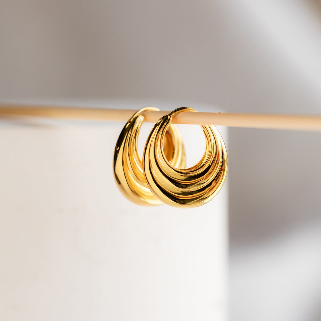 Two shiny Katrina Ribbed Hoops with a chunky, ribbed texture hang on a thin rod against a light, blurred background.