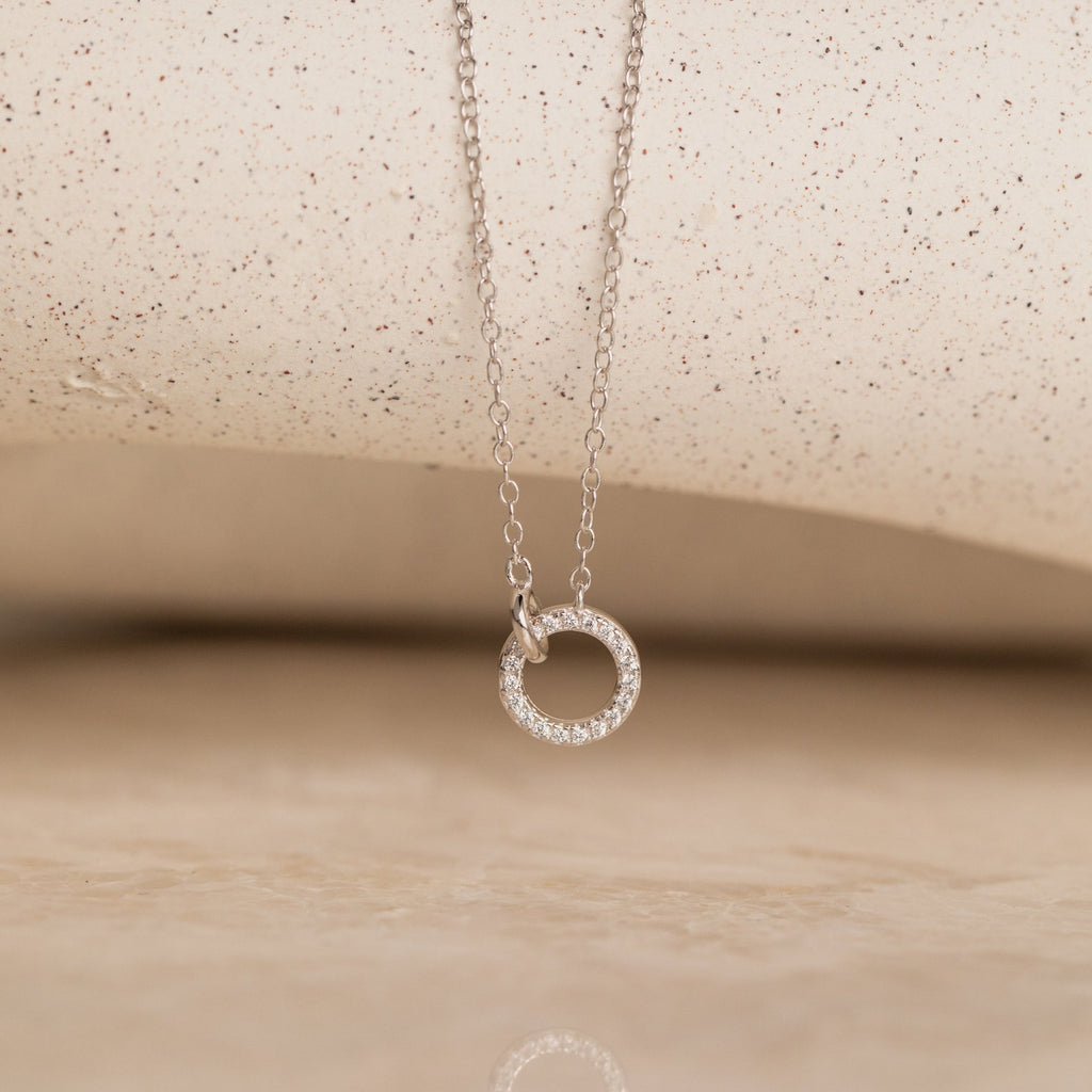The Pave Interlocking Necklace features a delicate silver chain with a diamond-encrusted open circle pendant, displayed on a neutral background—perfect for minimal diamond jewelry lovers or as a thoughtful wedding gift.