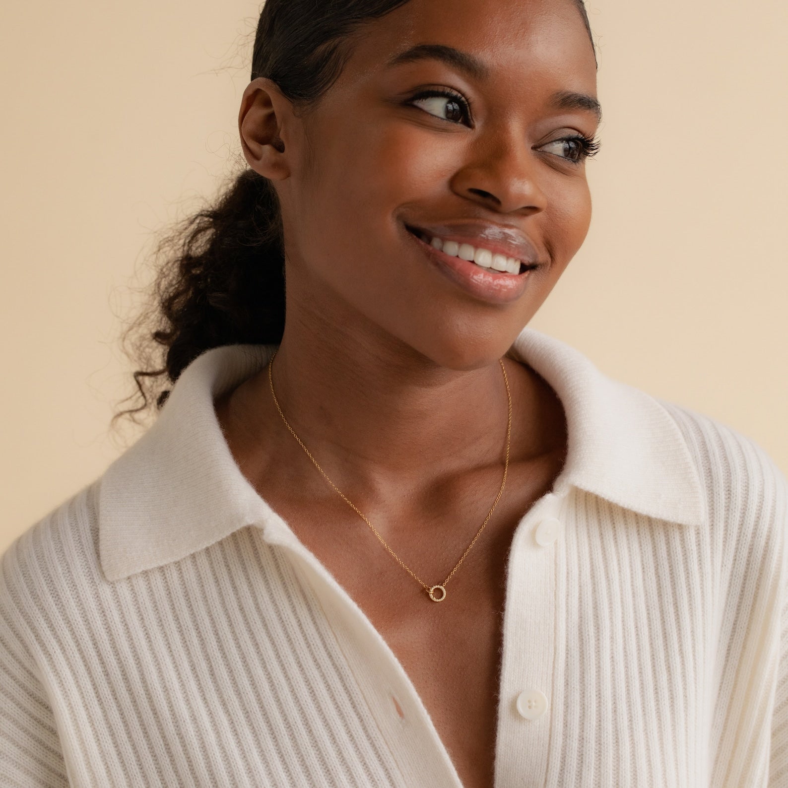 A woman smiles in a white ribbed sweater, wearing the Pave Interlocking Necklace—a dainty gold circle pendant—an elegant minimal diamond jewelry piece or perfect wedding gift, set against a beige background.