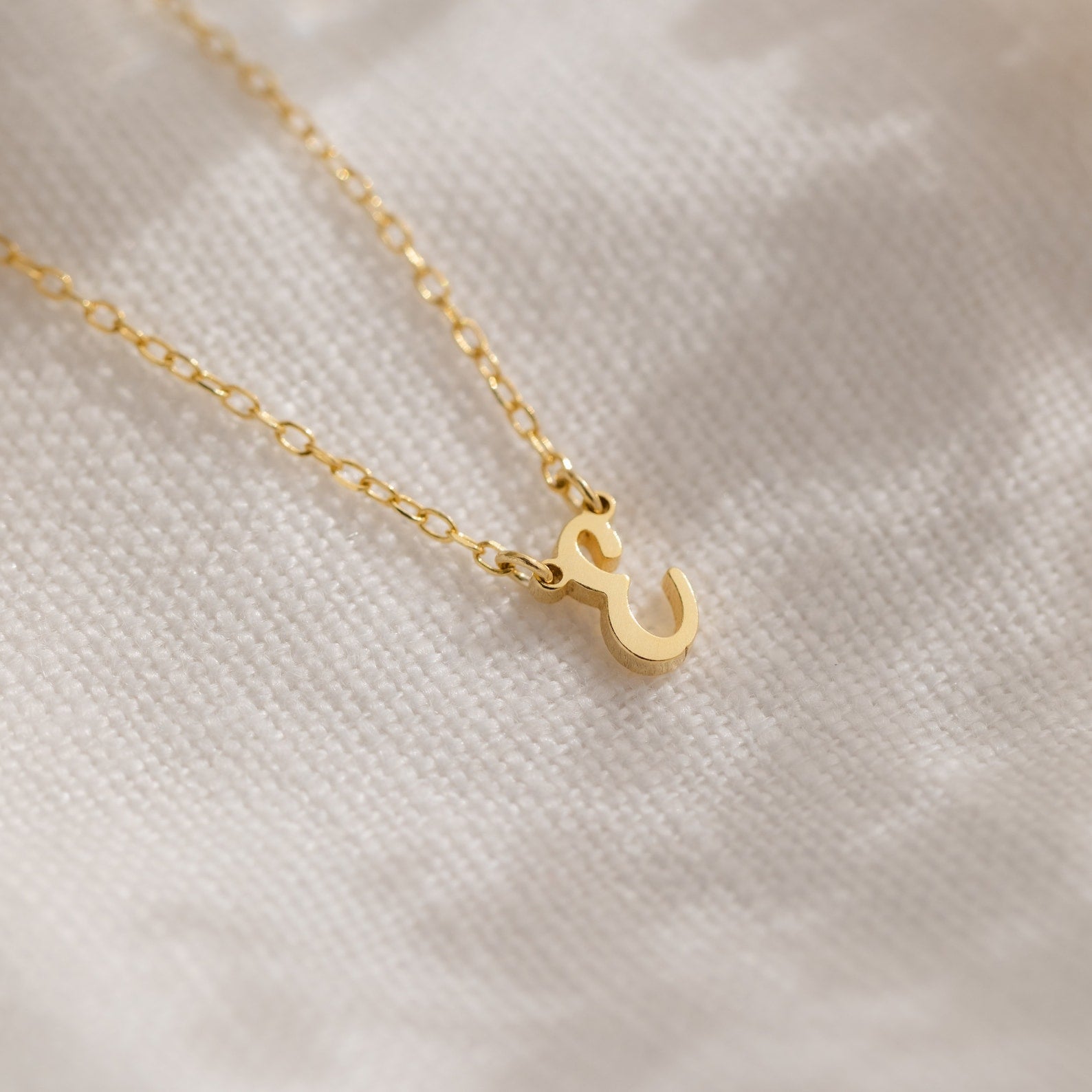 The Stylish Dainty Initial Necklace, featuring a gold ampersand pendant, is displayed on soft, white textured fabric.