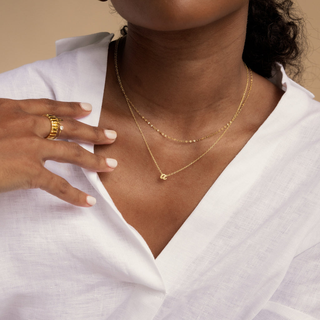 A woman in a white blouse wears layered gold necklaces, featuring the Grace Dainty Initial Necklace and a gold ring, as she gently touches her collarbone.
