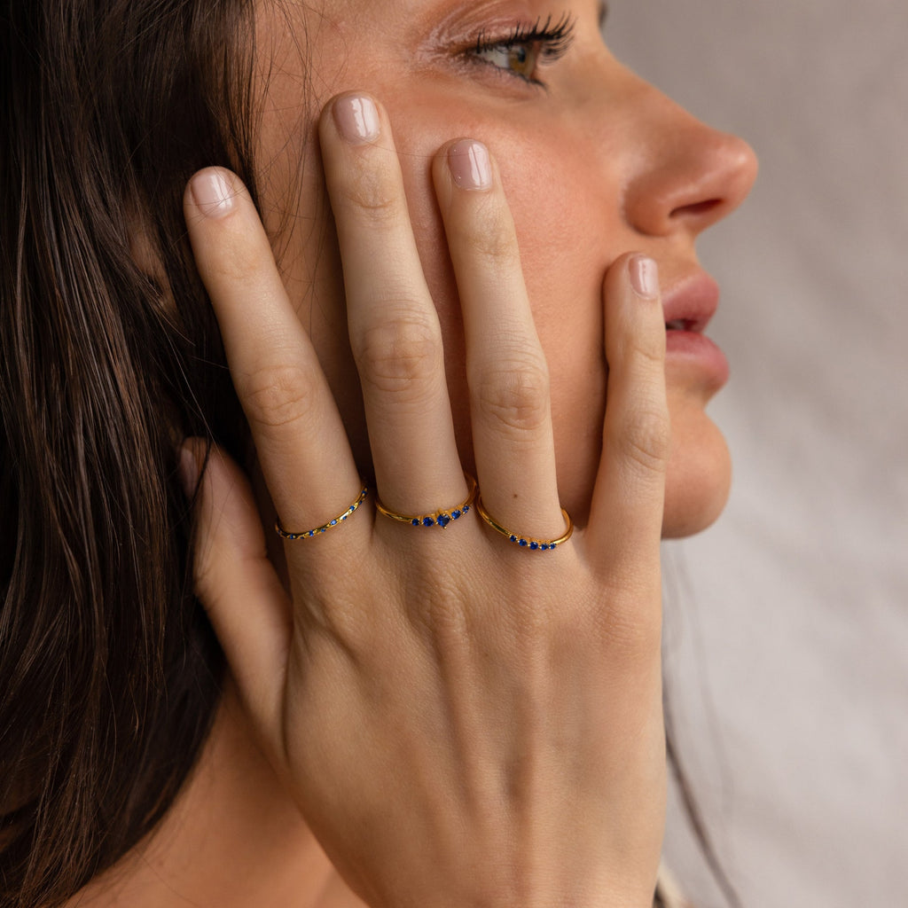 A woman with manicured nails touches her face, showcasing three gold rings, including the Sapphire Stacking Ring Set adorned with sparkling blue sapphires.