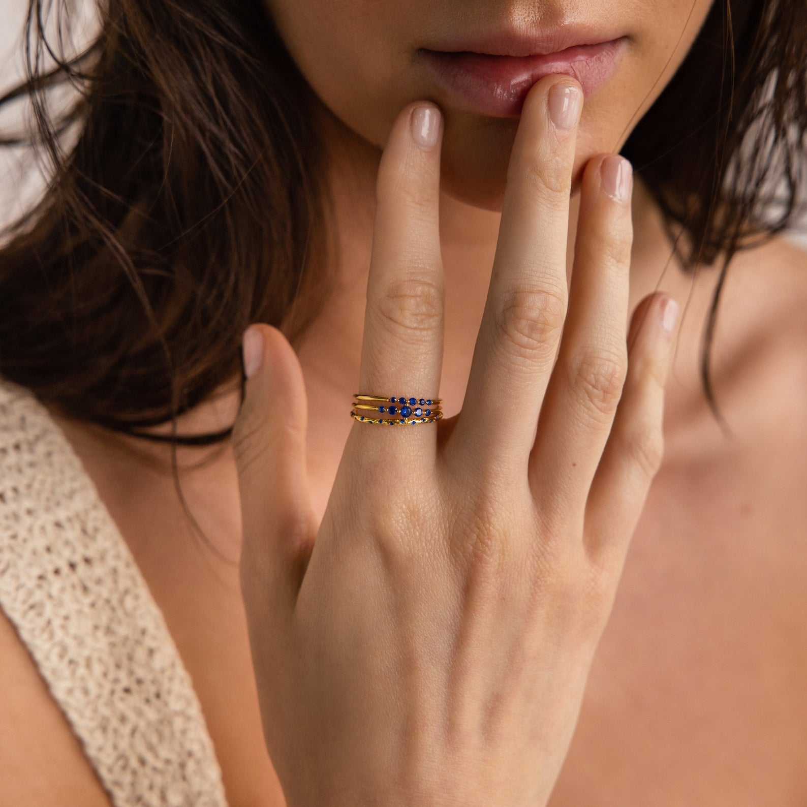 Close-up of a hand wearing a set of 3 gold rings with blue stones, brushing against the lips.