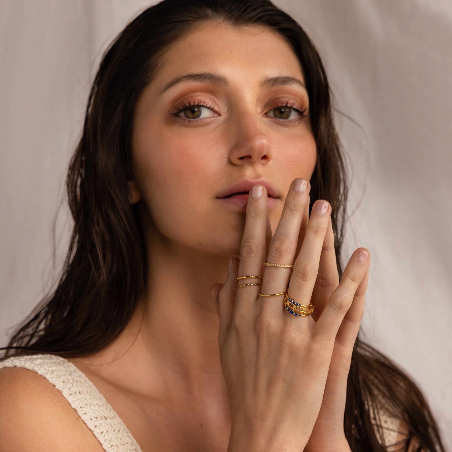 Woman with hand near face wearing various gold rings on a neutral background
