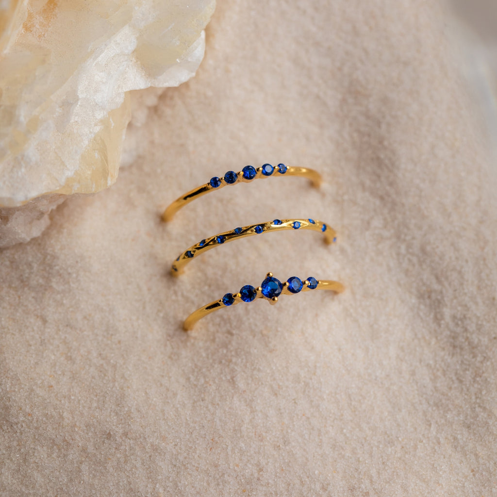 The Sapphire Stacking Ring Set features three gold rings with blue sapphires, elegantly displayed on light sand next to a translucent crystal.