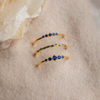 The Sapphire Stacking Ring Set features three gold rings with blue sapphires, elegantly displayed on light sand next to a translucent crystal.