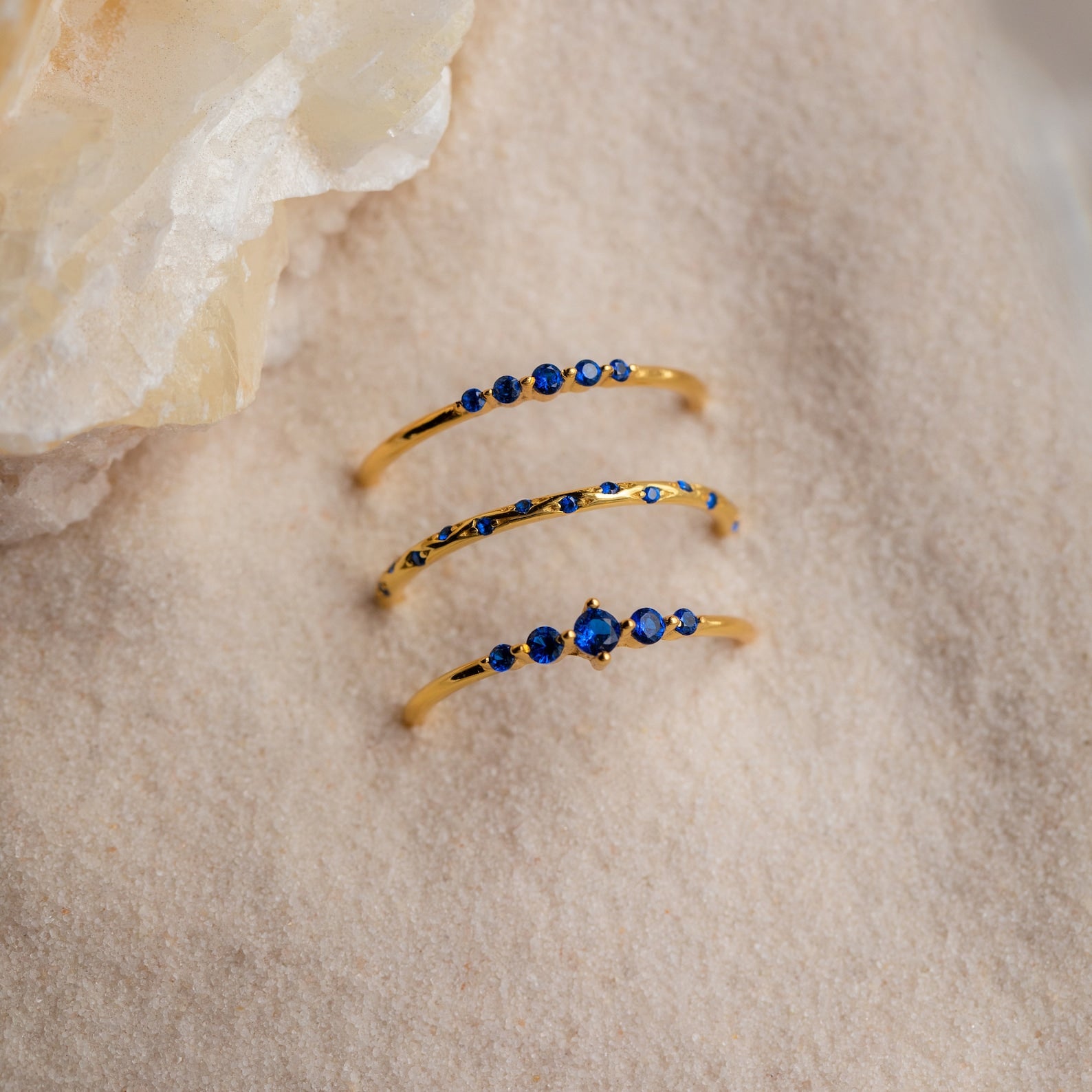 Three gold rings with blue stones on a textured sandy surface