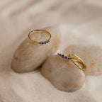 The Sapphire Stacking Ring Set features two gold rings with small blue sapphires, displayed on smooth rocks surrounded by sand.
