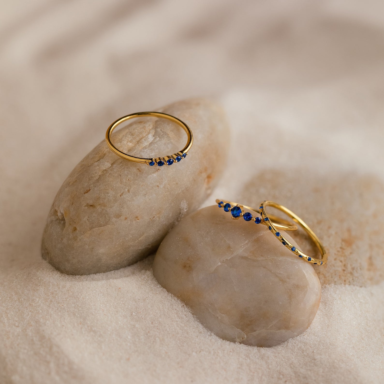 The Sapphire Stacking Ring Set features two gold rings with small blue sapphires, displayed on smooth rocks surrounded by sand.