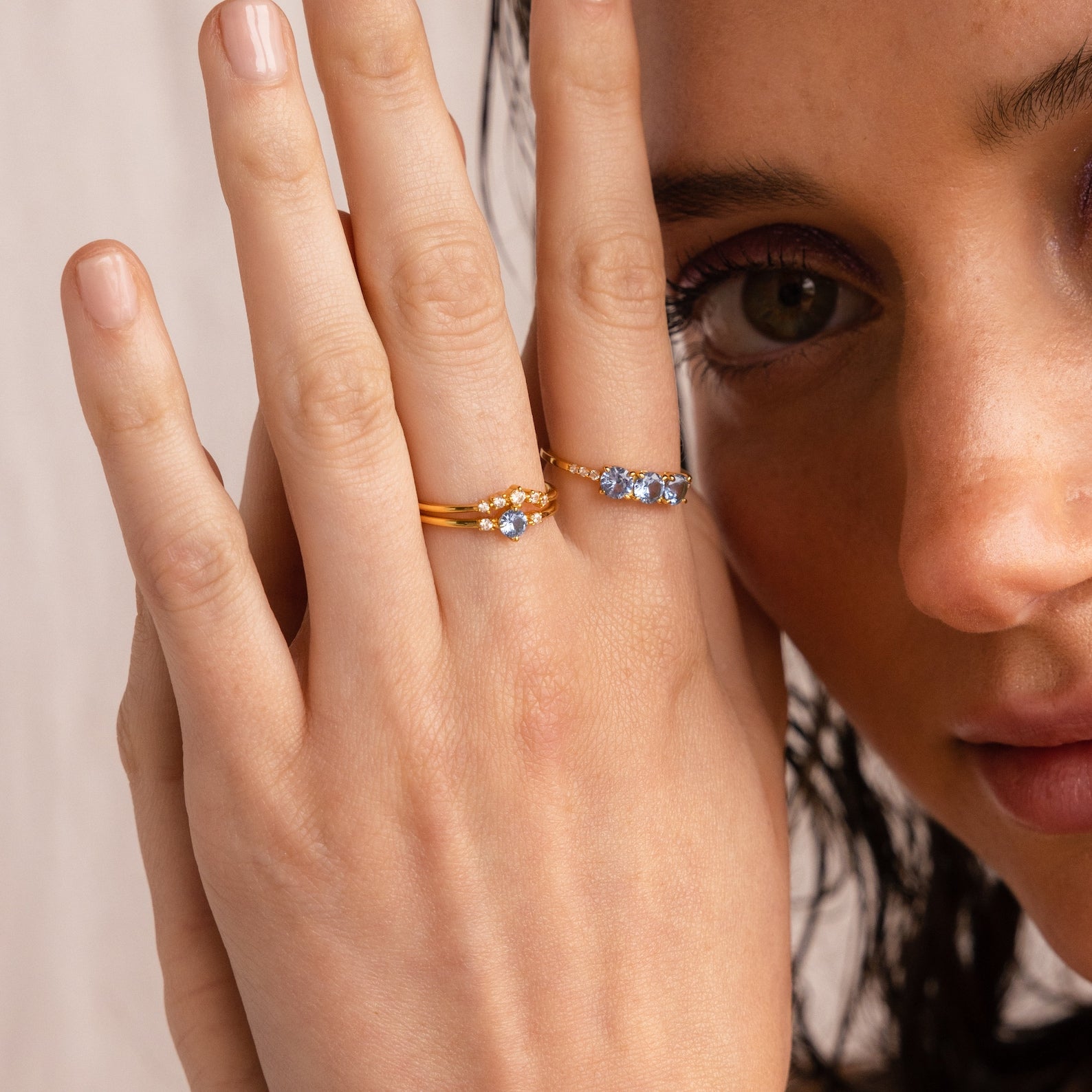 A woman with light skin wears the Aquamarine Stacking Ring Set, featuring gold bands with blue and white stones, on her fingers near her face.