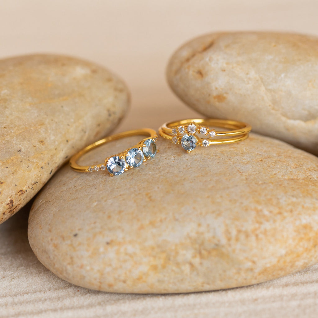 The Aquamarine Stacking Ring Set features two gold boho rings—one with sparkling ocean blue gemstones and the other an elegant aquamarine band—displayed on smooth, light-colored stones.