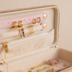 The Rectangle Velvet Jewelry Case in pink velvet is open, showing gold earrings, a zippered pocket, and gold necklaces arranged against a beige background.