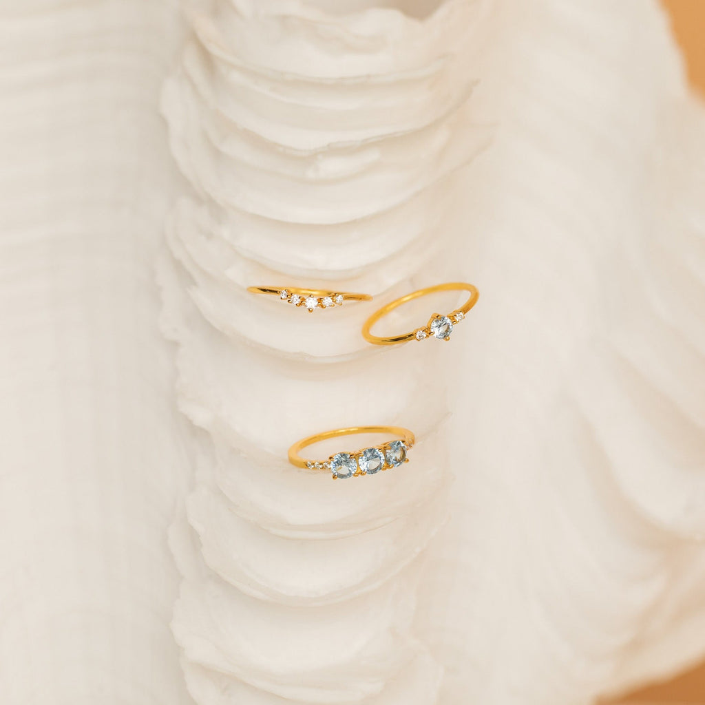 The Aquamarine Stacking Ring Set features three delicate gold boho rings with ocean blue gemstones, beautifully displayed on a textured white seashell.