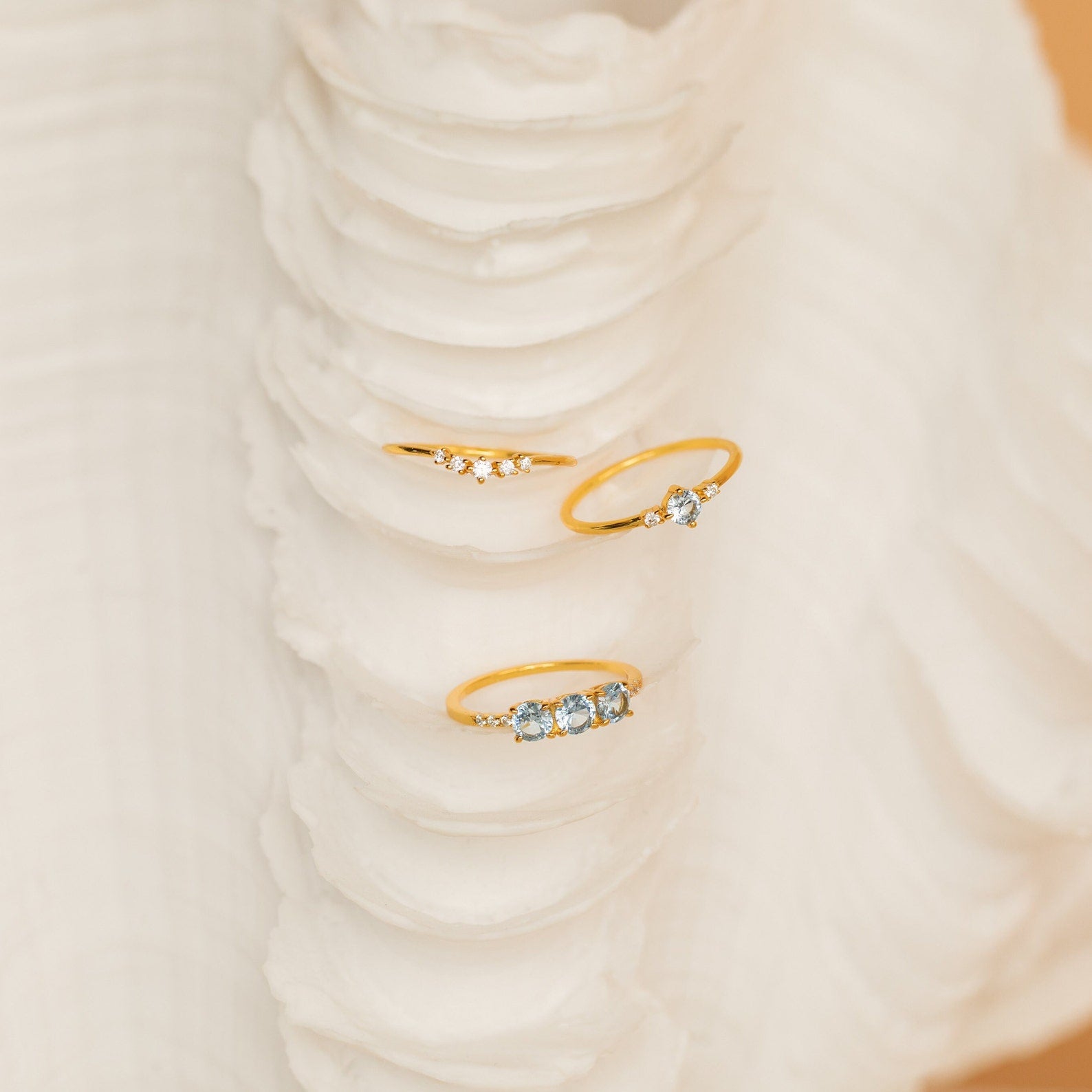 The Aquamarine Stacking Ring Set features three delicate gold boho rings with ocean blue gemstones, beautifully displayed on a textured white seashell.