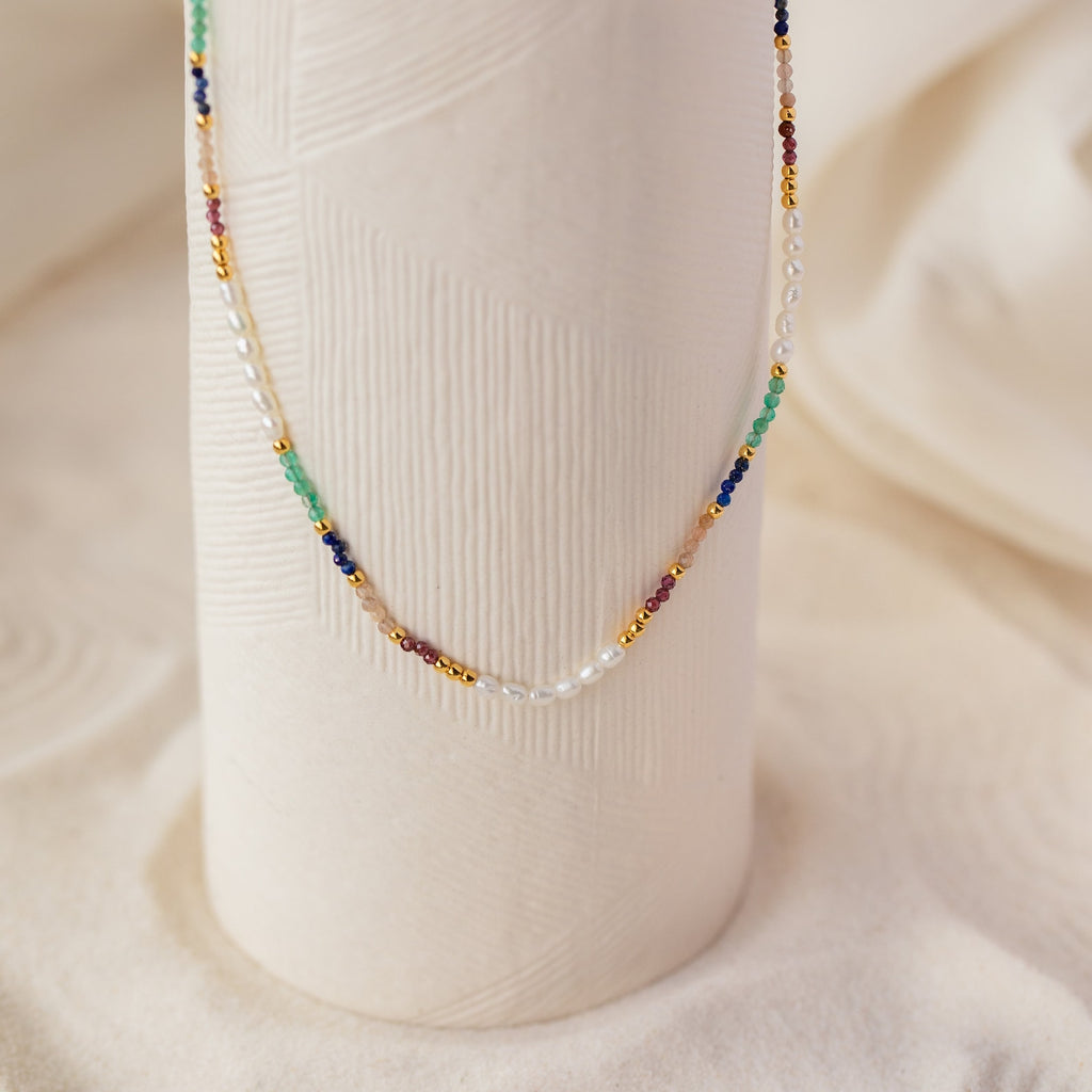 The Oasis Rainbow Beaded Necklace, featuring small pearls and colorful beads, drapes elegantly over a textured white vase—making it the perfect boho jewelry or summer accessory.